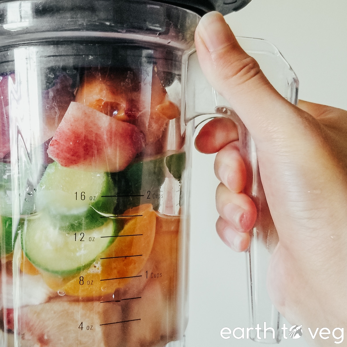 A hand holds up a Blendtec Twister Jar full of zucchini, peach, and nectarine.