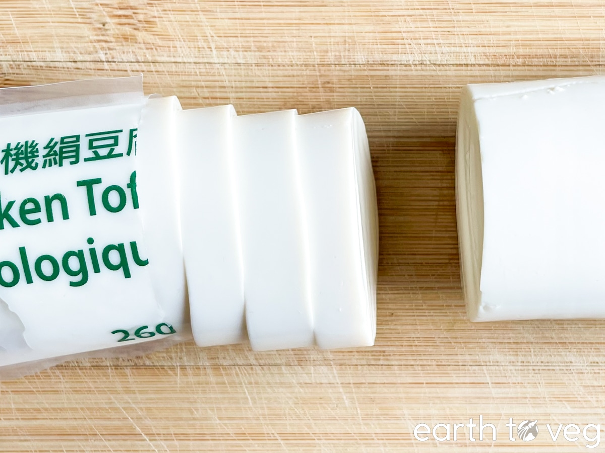 A tube of silken tofu, partially sliced, on a wooden cutting board.