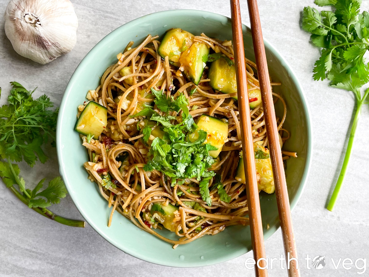 Bean curd cucumber salad in a teal green bowl with a pair of wooden chopsticks surrounded by fresh garlic and herbs.