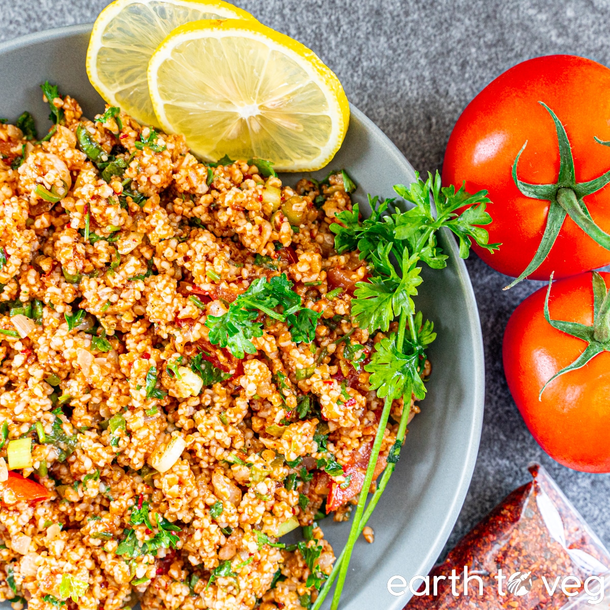 A grey bowl filled with kisir salad is garnished with parsley sprigs, lemon slices, and tomatoes.