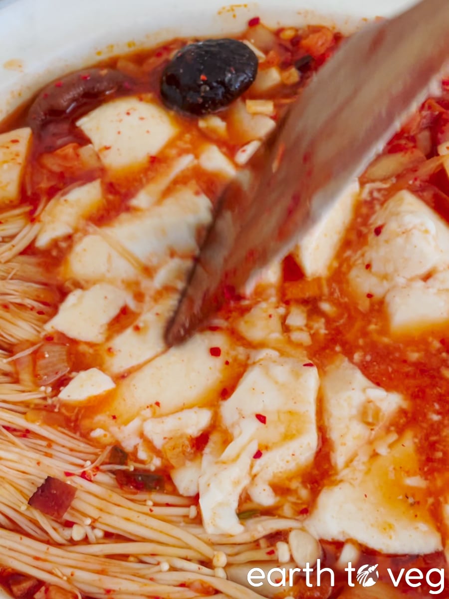 Silken tofu has been added to the pot and is being gently broken up by a wooden spatula.
