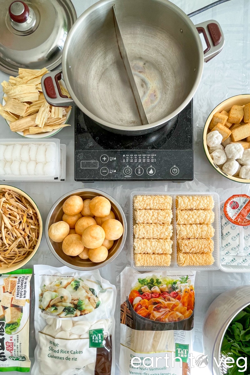 A top-down view of various Chinese hot pot ingredients laid out on a dining table, with a yuanyang split hot pot on an induction stove in the center of the table.