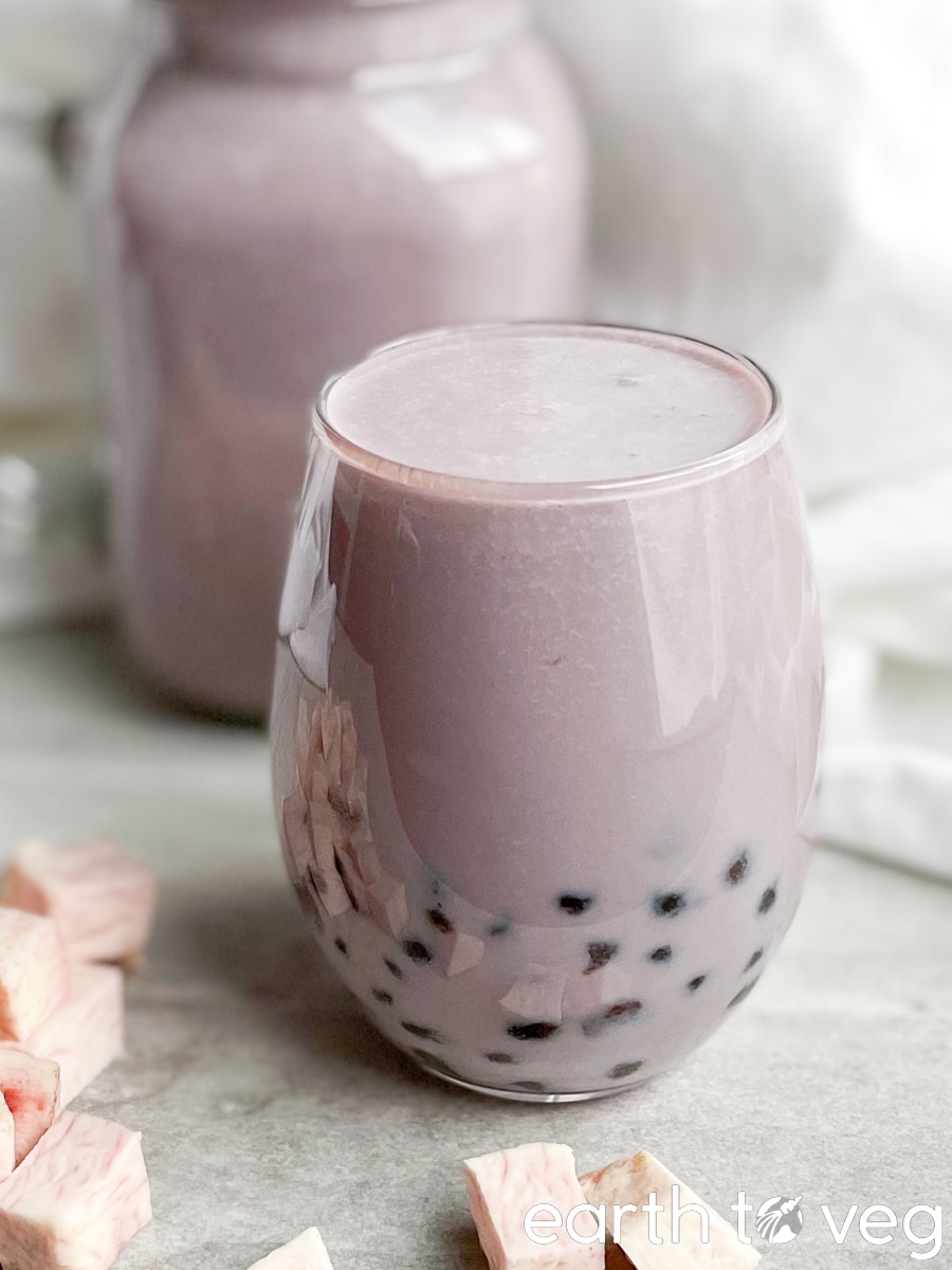 A round glass of taro milk tea filled with tapioca pearls is stood in front of a mason jar glass full of the same drink, in the background.