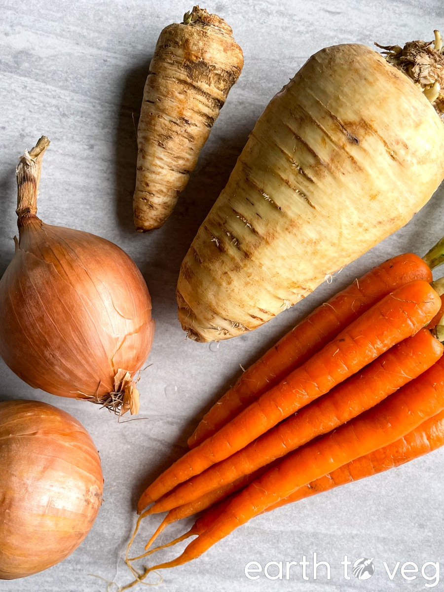 Unpeeled assortment of onions, carrots, and parsnips on a grey countertop.