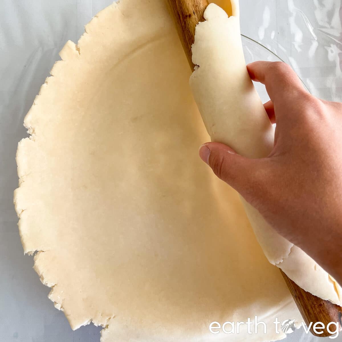 A hand holding a rolling pin laying out a pie crust over a glass pie pan.