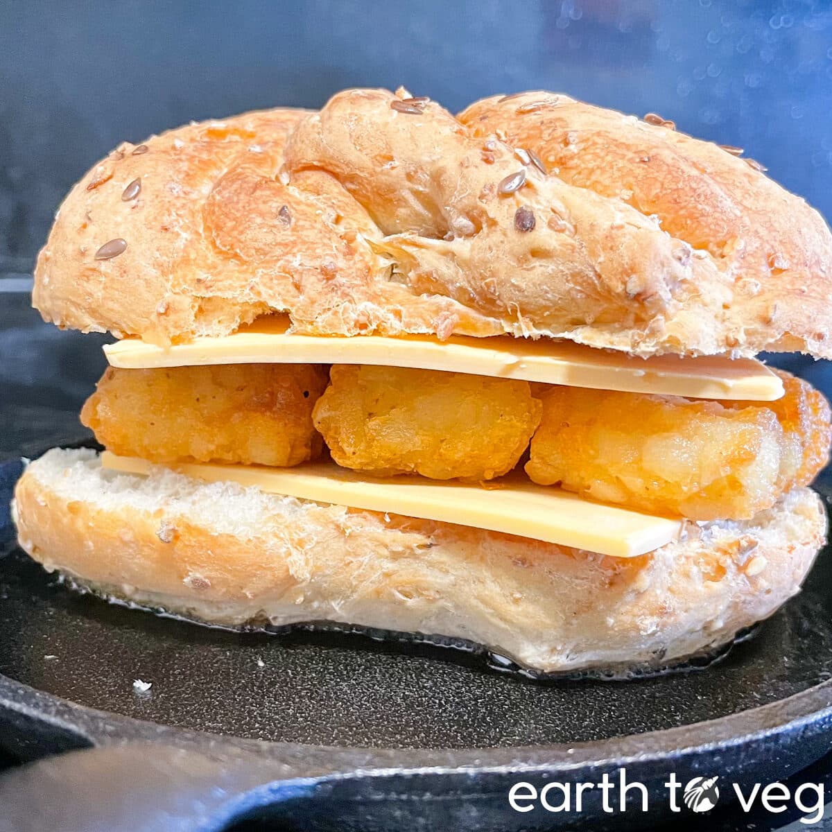assembled but not yet cooked tater tot sandwich with kaiser roll, vegan cheddar cheese, and tater tots, on a cast iron pan