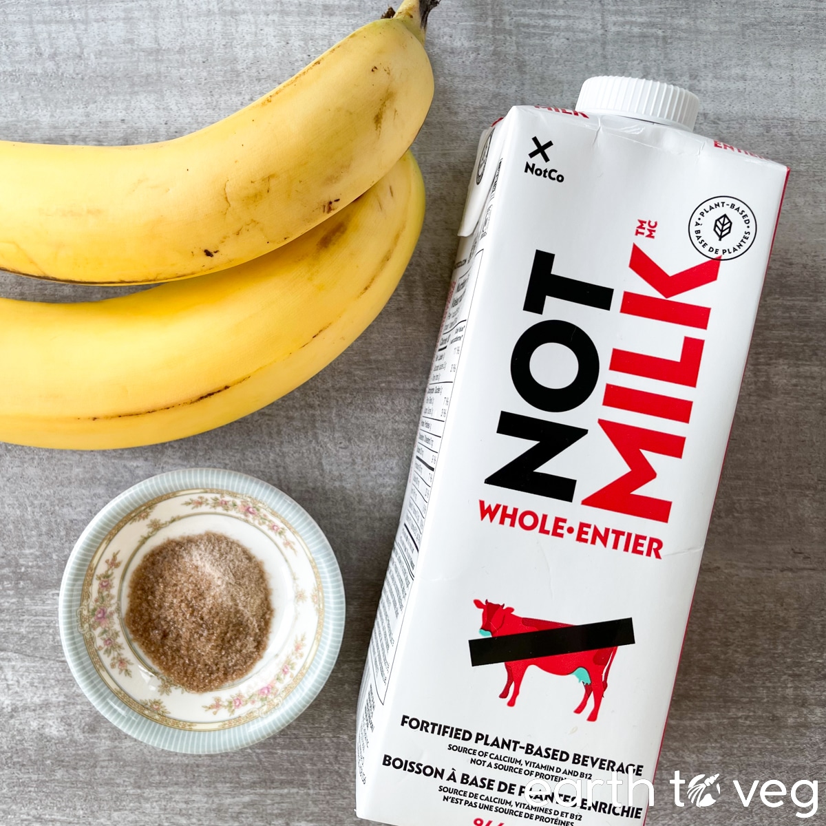A top-down view of the ingredients for a banana licuado smoothie: two bananas, a bowl of brown sugar, and a carton of NotMilk plant-based beverage are on a grey countertop.
