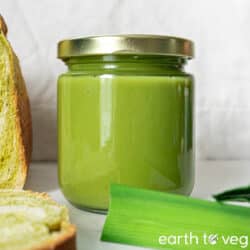 A jar of vegan pandan kaya sits on a white table next to fresh pandan leaves and a loaf of pandan bread.