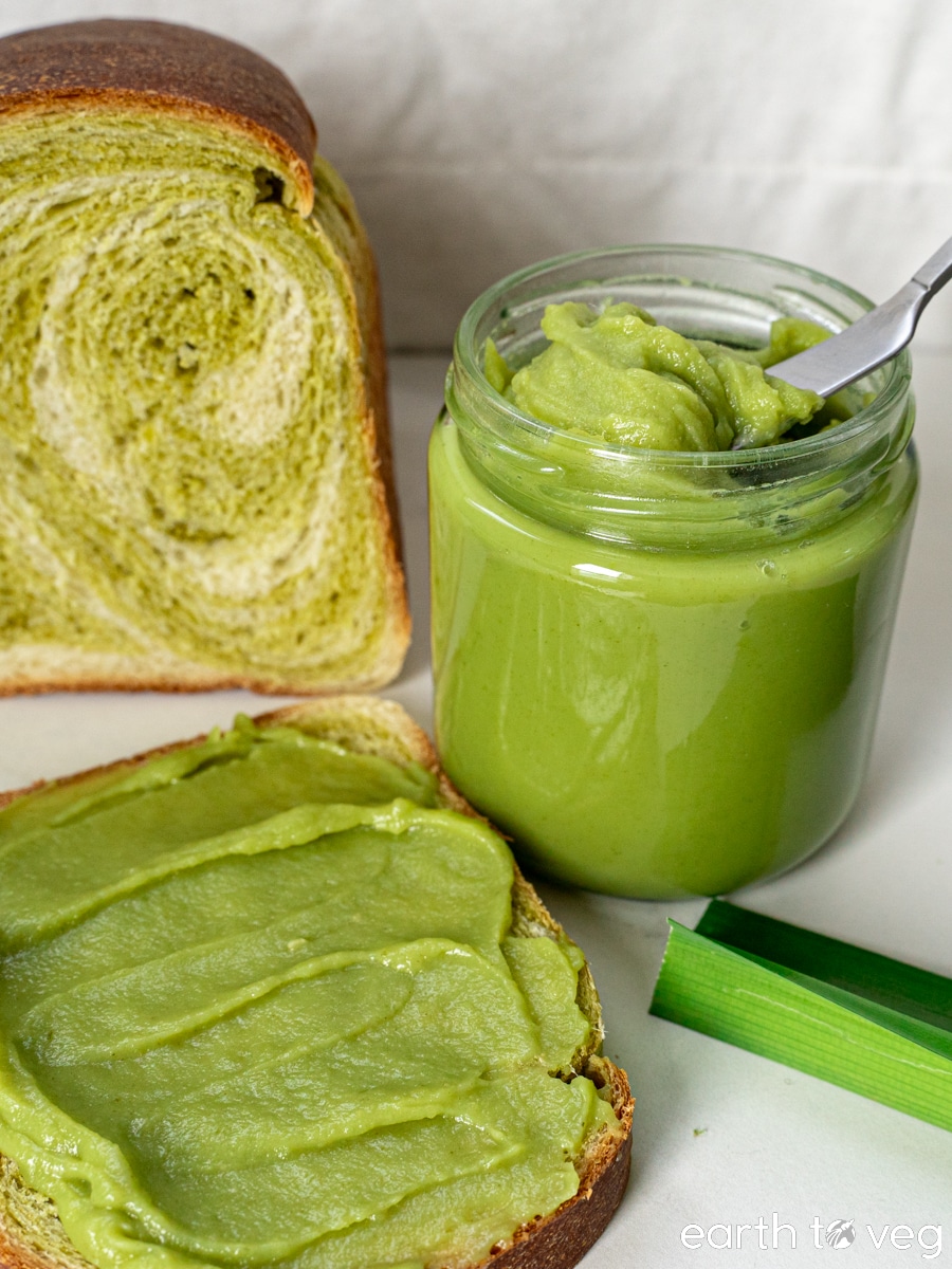A jar of Nyonya kaya sits on a white table next to fresh pandan leaves and a loaf of pandan bread.