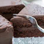 A metal fork cuts into a double-layer chocolate cake with a thick layer of chocolate frosting.