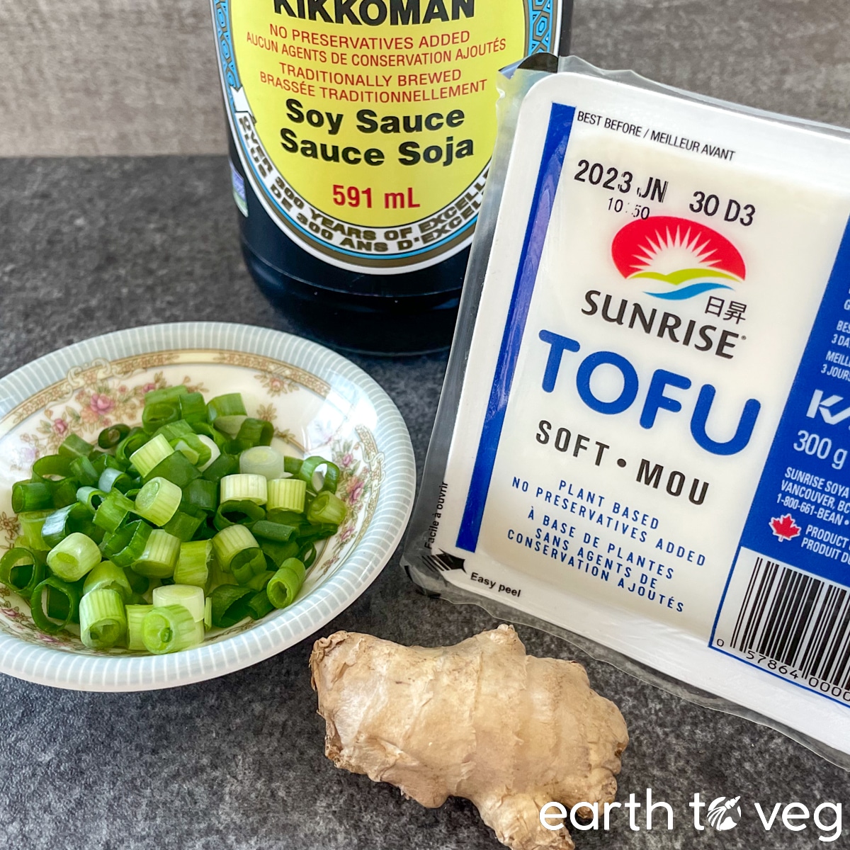 A box of soft tofu on the countertop, next to freshly diced scallions, a small thumb of ginger, and a bottle of soy sauce.
