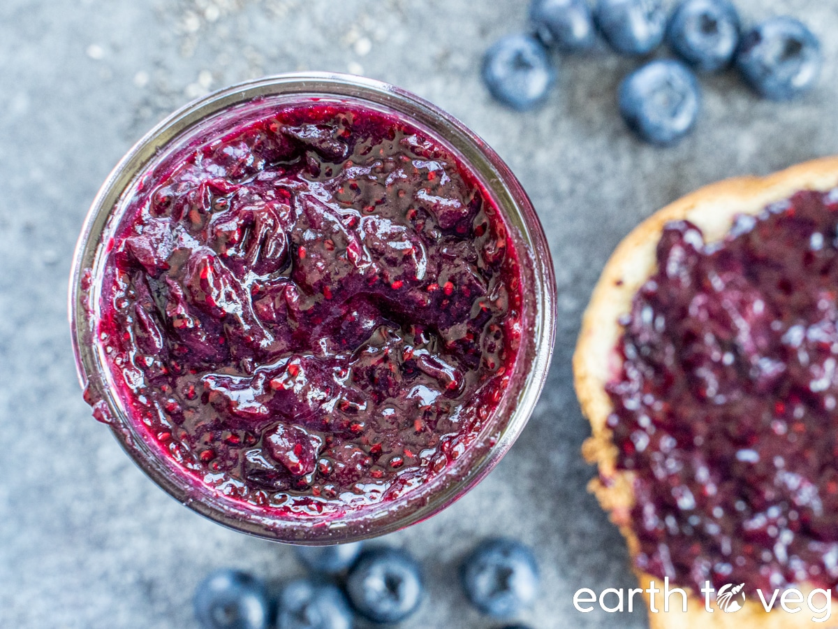 Blueberry chia jam on a grey countertop next to a slice of toast spread with jam and some scattered fresh blueberries.