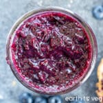 A glass pot filled to the brim with blueberry chia jam, on a grey-blue countertop surrounded by fresh blueberries.
