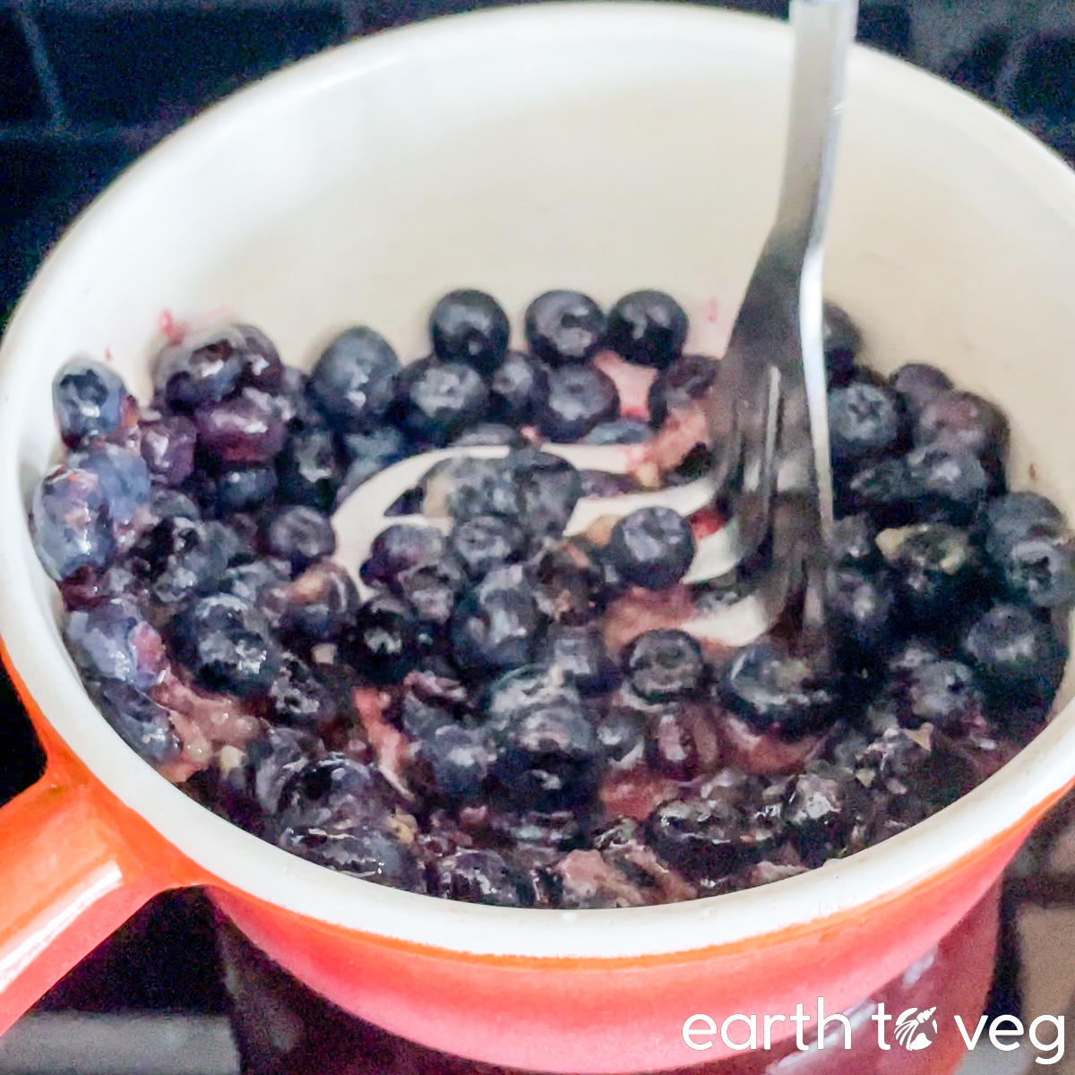 Using a potato masher to break down blueberries in a small ceramic pot.