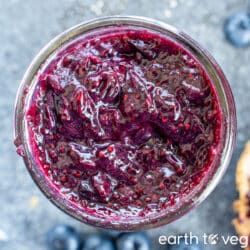 A glass pot filled to the brim with blueberry chia jam, on a grey-blue countertop surrounded by fresh blueberries.