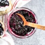 A copper spoon rests in a jar of haskap jam, next to a tea towel on which rests two scones with cream and jam.