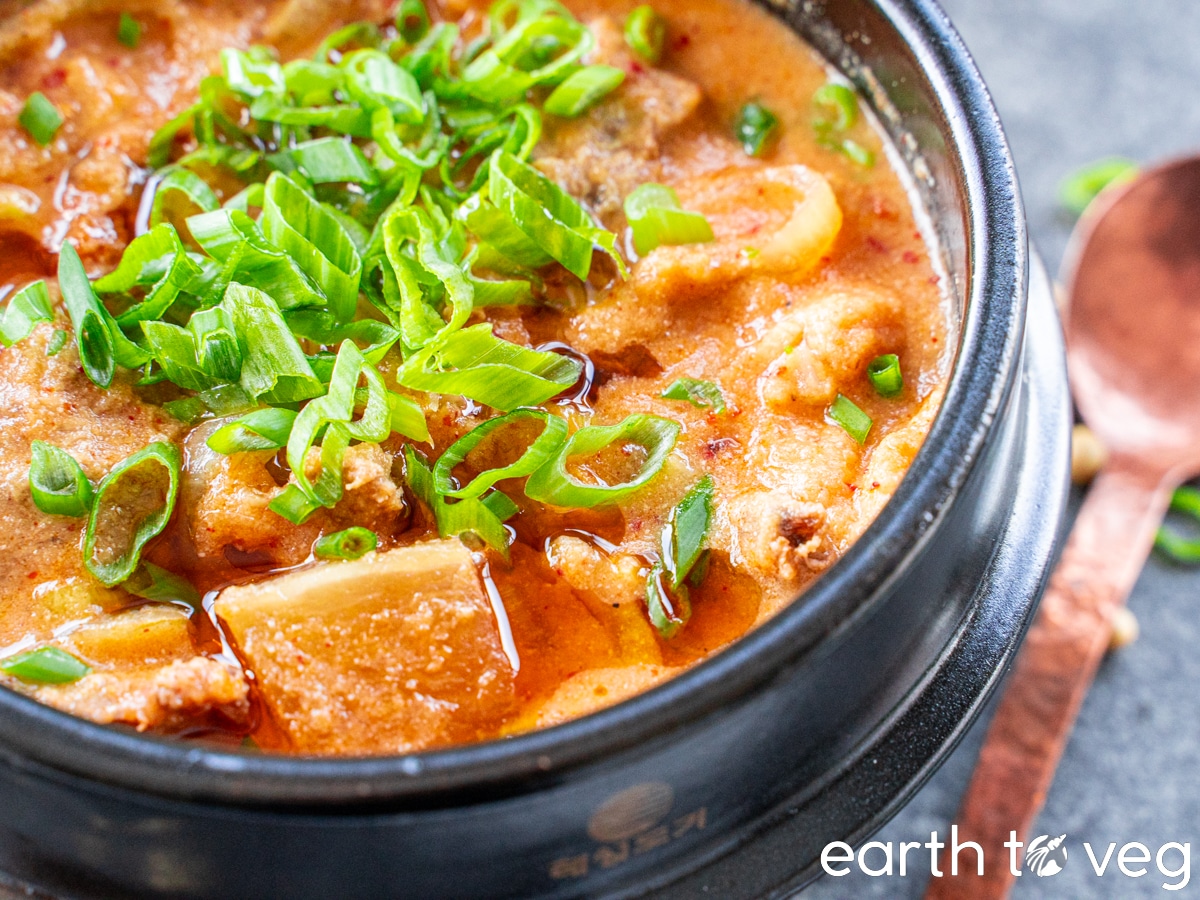 A ddukbaegi filled with Korean kimchi okara stew strewn with scallions and sesame oil, on a grey countertop next to a copper spoon.