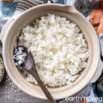 A donabe full of cooked white rice with a dark wooden spoon laid on top, on a grey counter next to colourful fabric.
