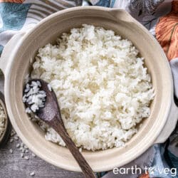 A donabe full of cooked white rice with a dark wooden spoon laid on top, on a grey counter next to colourful fabric.