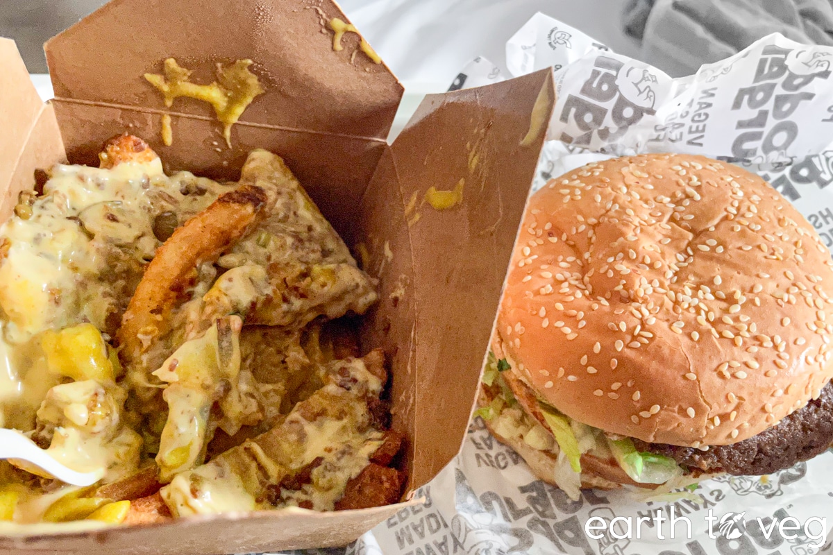 A paper takeout box of Famous-Style Fries next to a Famous Burger from Odd Burger.