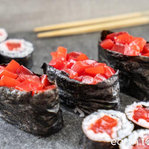 Three pieces of vegan gunkan maki are flanked by vegan maki rolls on a grey surface, with chopsticks in the background.