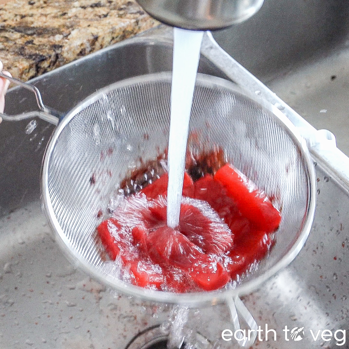 Red-dyed vegan sashimi is rinsed under running tap water in a metal colander.