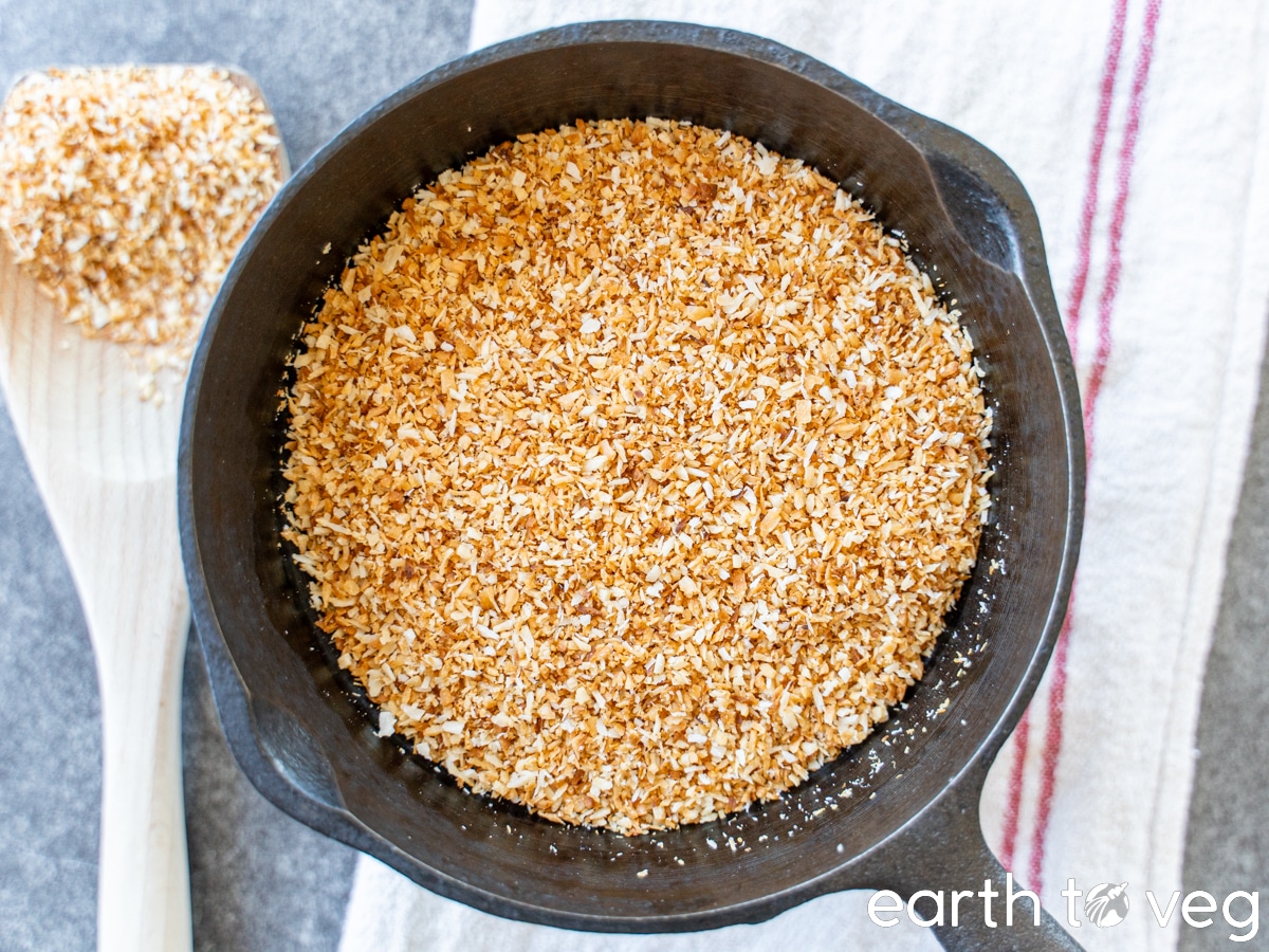 Toasted coconut shreds in a small cast iron pan on a white linen towel and next to a wooden spatula filled with more coconut.
