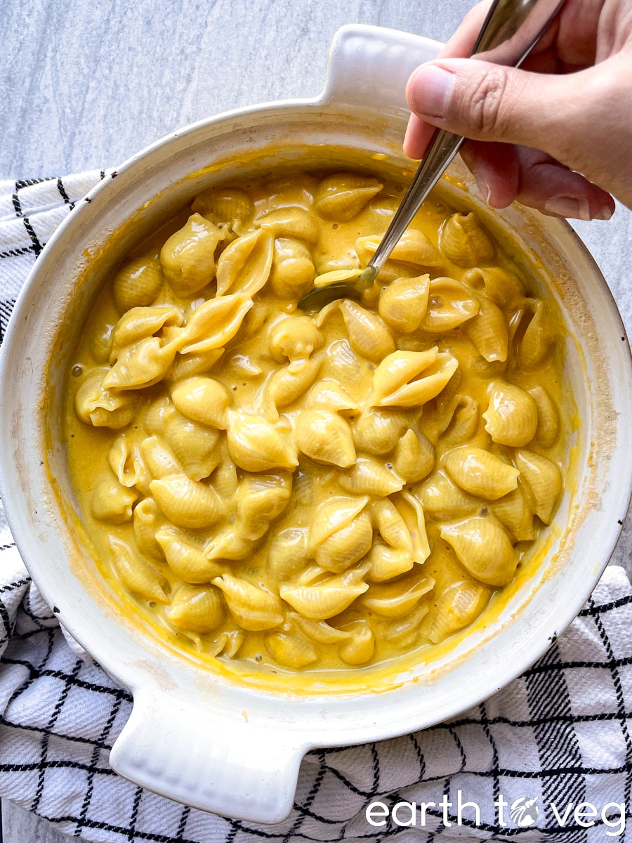 A hand stirs a spoon through a Dutch oven filled with gooey, saucy vegan macaroni and cheese.
