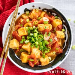 Chopped green scallions scattered over a bowl of tofu puffs smothered in tomato sauce, with a golden pair of chopsticks.