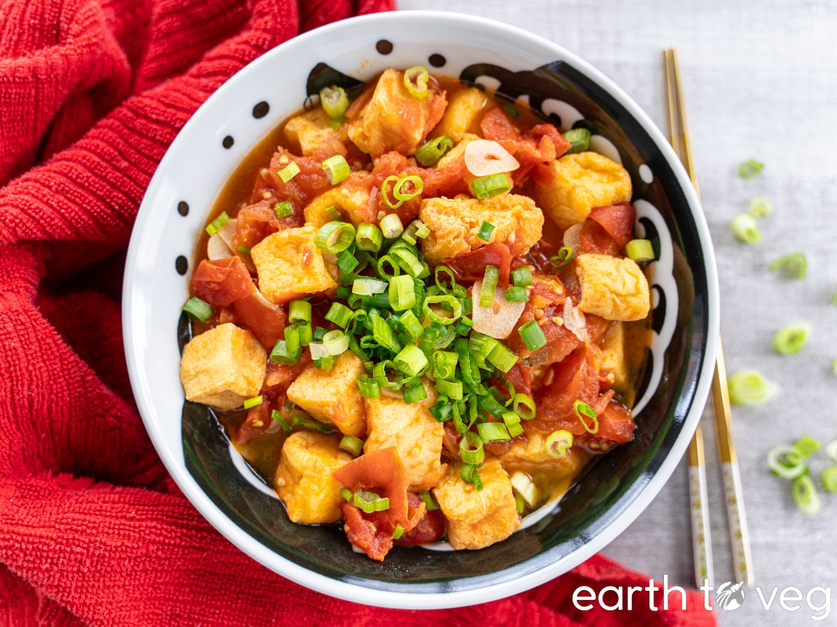 A large black-and-white bowl of Vietnamese tomato sauce with fried tofu on a smooth grey counter, next to a red towel and pair of chopsticks.