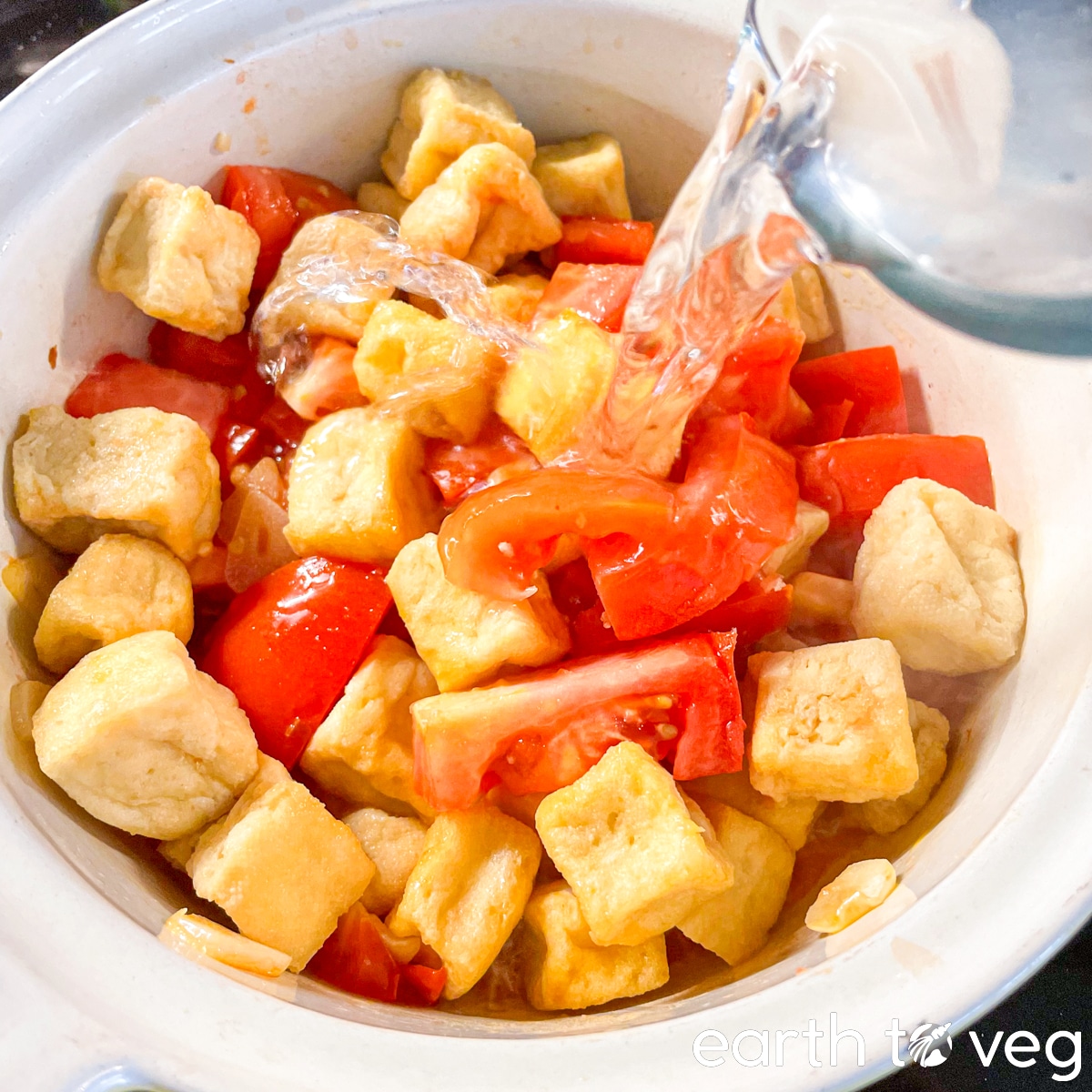 Water is poured from a glass measuring cup into a ceramic Dutch oven full of tofu puffs and fresh tomatoes.