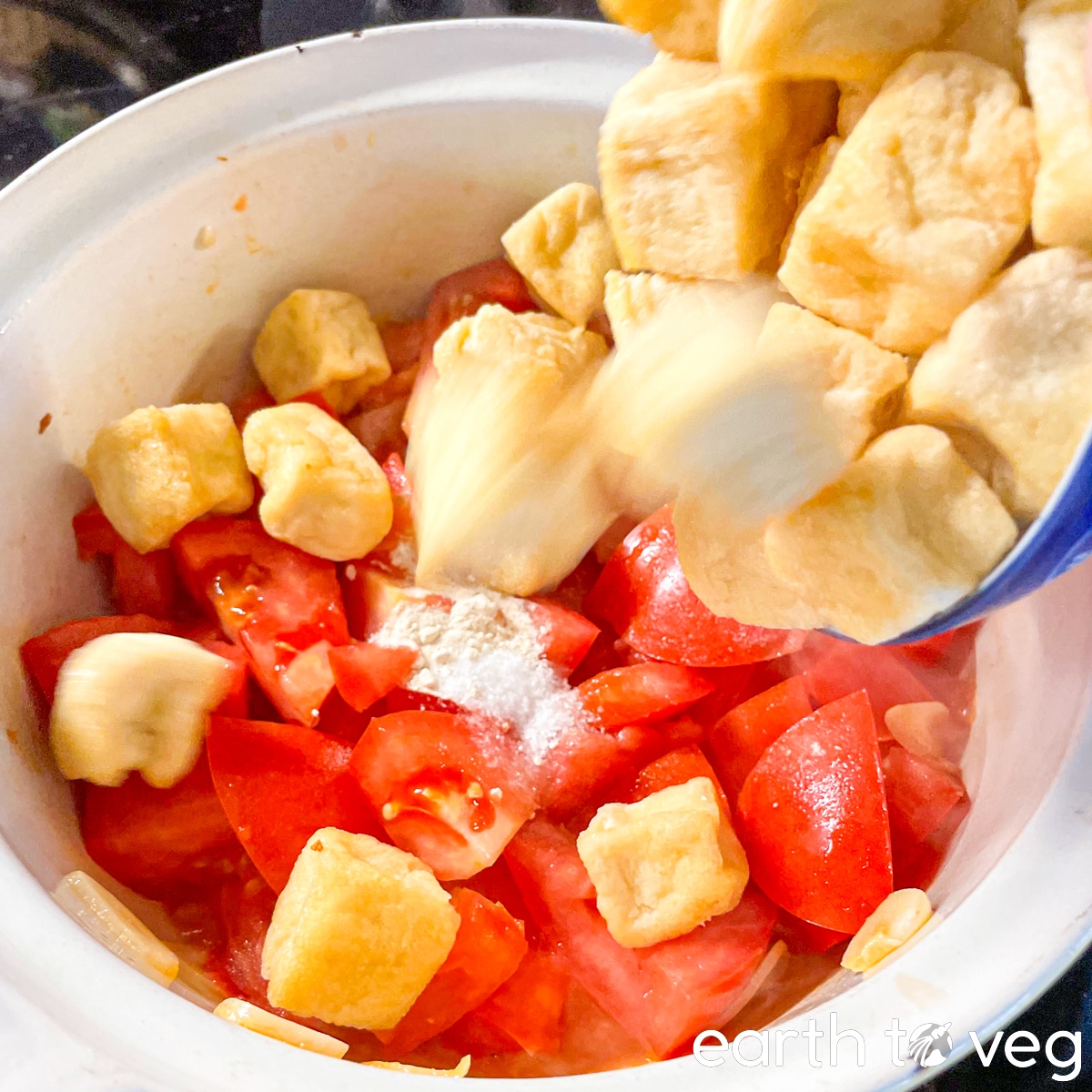 Tofu puffs and seasonings tumble from a bowl into a pot full of chopped tomatoes.