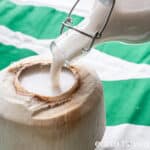 Coconut milk is poured from a glass bottle into a hollowed-out coconut shell.