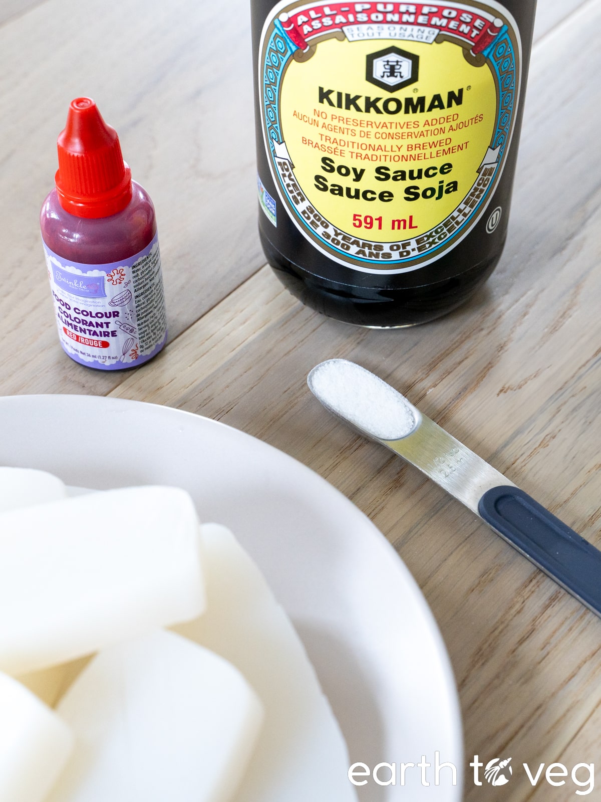 A bottle of soy sauce, a plate of konjac slices, a spoon of sugar, and a small jar of red food colouring on a wooden surface.