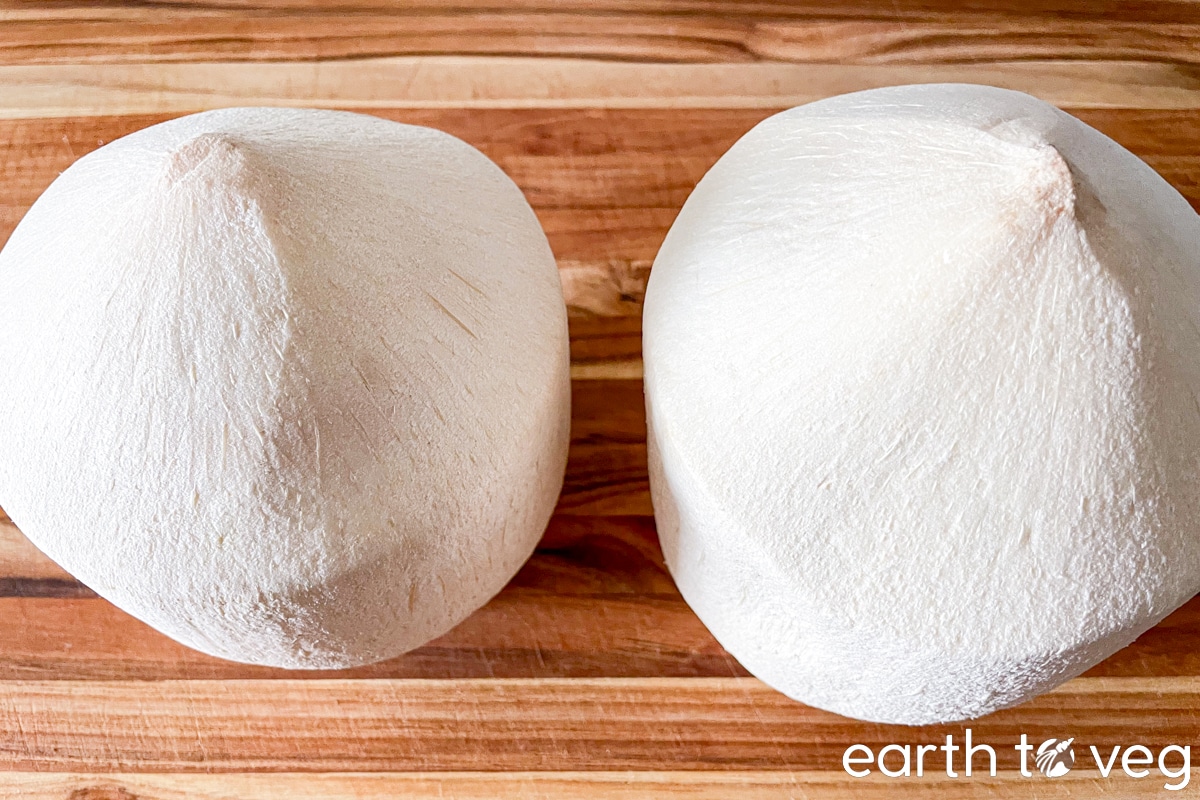 Two husked young coconuts side by side on a wooden cutting board.