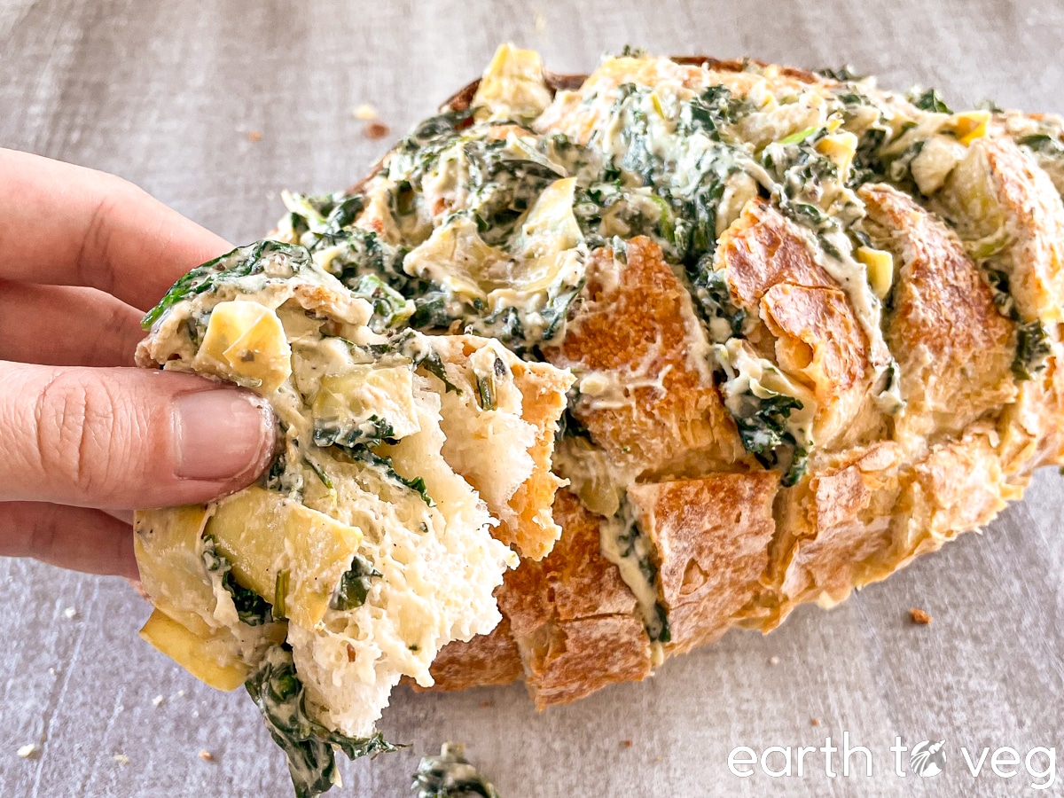 A hand holds up a torn piece of spinach artichoke pull-apart bread.