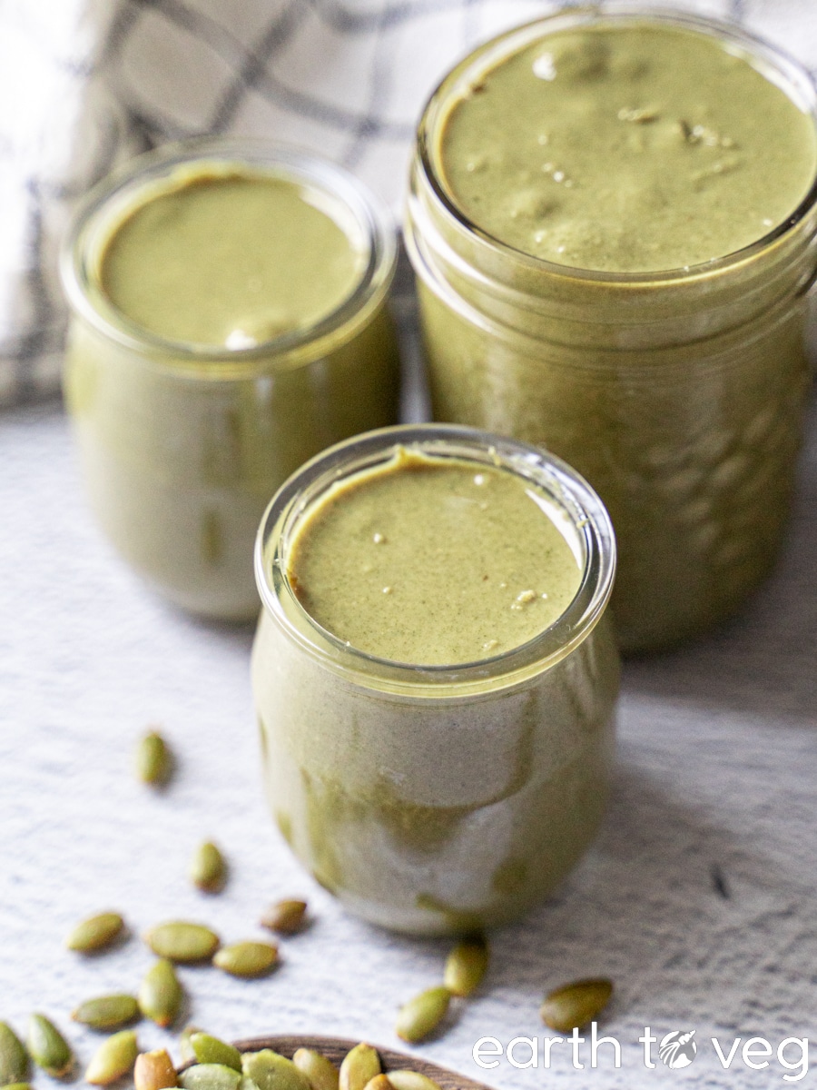 Three jars full of roasted pumpkin seed butter on a grey table, with scattered pepitas nearby.