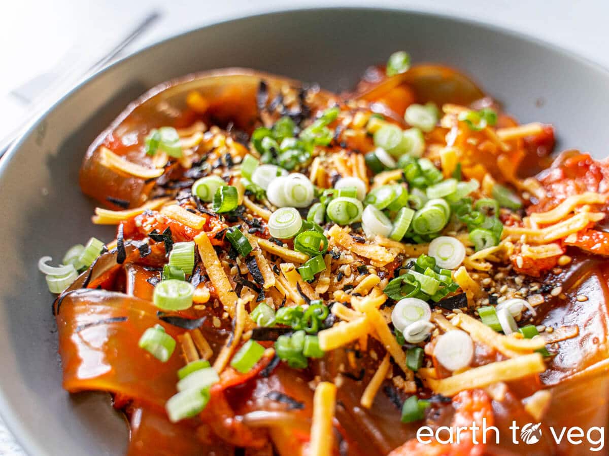 Spicy Korean sweet potato noodles coated with buldak hot sauce on a plate with scallions and shredded cheese.
