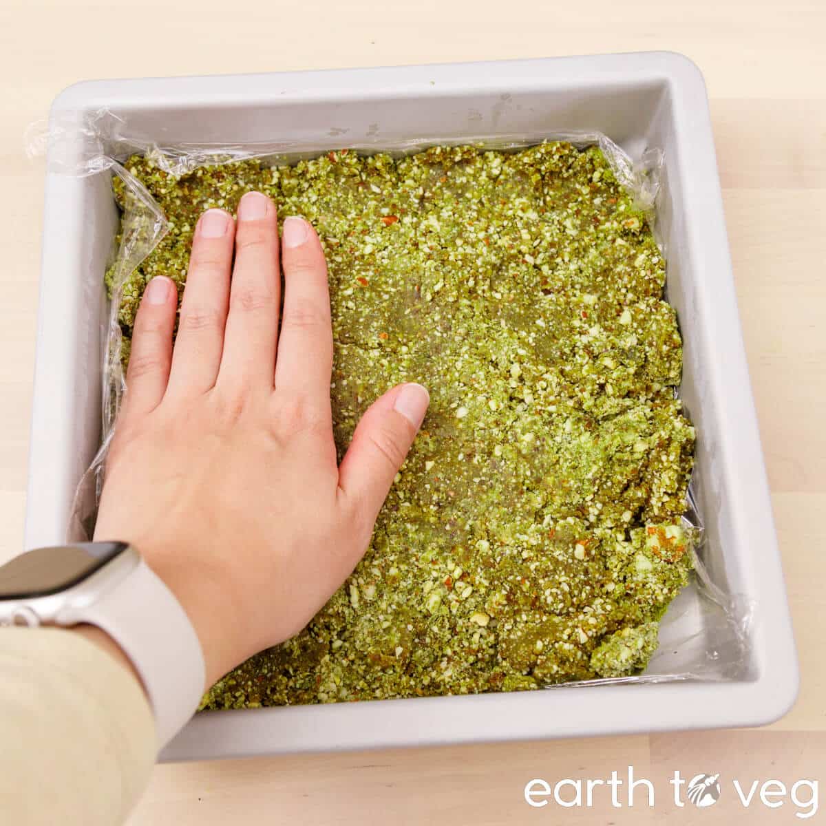 A hand presses matcha date dough into a square baking pan mold.