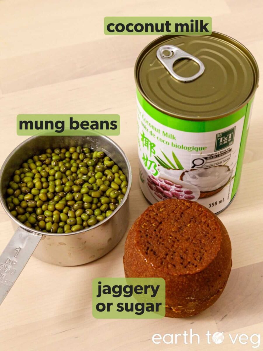 The three ingredients for godshe labelled on a wood surface: a cup of whole green mung beans, a can of coconut milk, and a round block of coconut palm sugar.