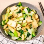 Marinated tofu planks tossed with crisp bok choy stems and dark green leaves in a grey bowl, glossy with sesame oil, chopsticks resting beside it.