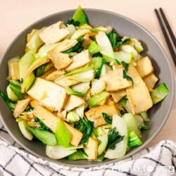 Marinated tofu planks tossed with crisp bok choy stems and dark green leaves in a grey bowl, glossy with sesame oil, chopsticks resting beside it.
