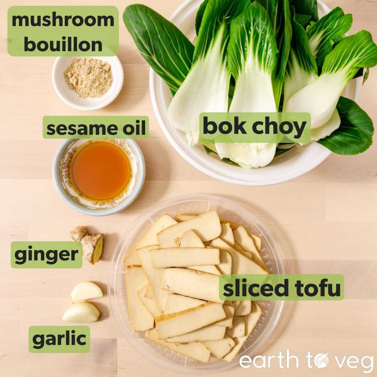 Labelled ingredients for bok choy tofu stir fry: a small bowl of mushroom bouillon powder, a saucer of sesame oil, baby bok choy, fresh ginger, two cloves of garlic, and a container of sliced marinated tofu.