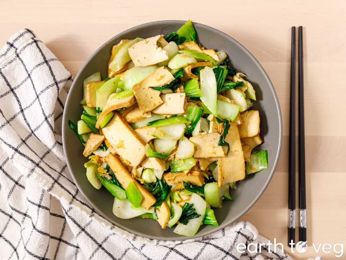 A bowl of bok choy tofu stir fry with marinated tofu planks and tender baby bok choy, set on a wooden table with black chopsticks and a checkered linen napkin.