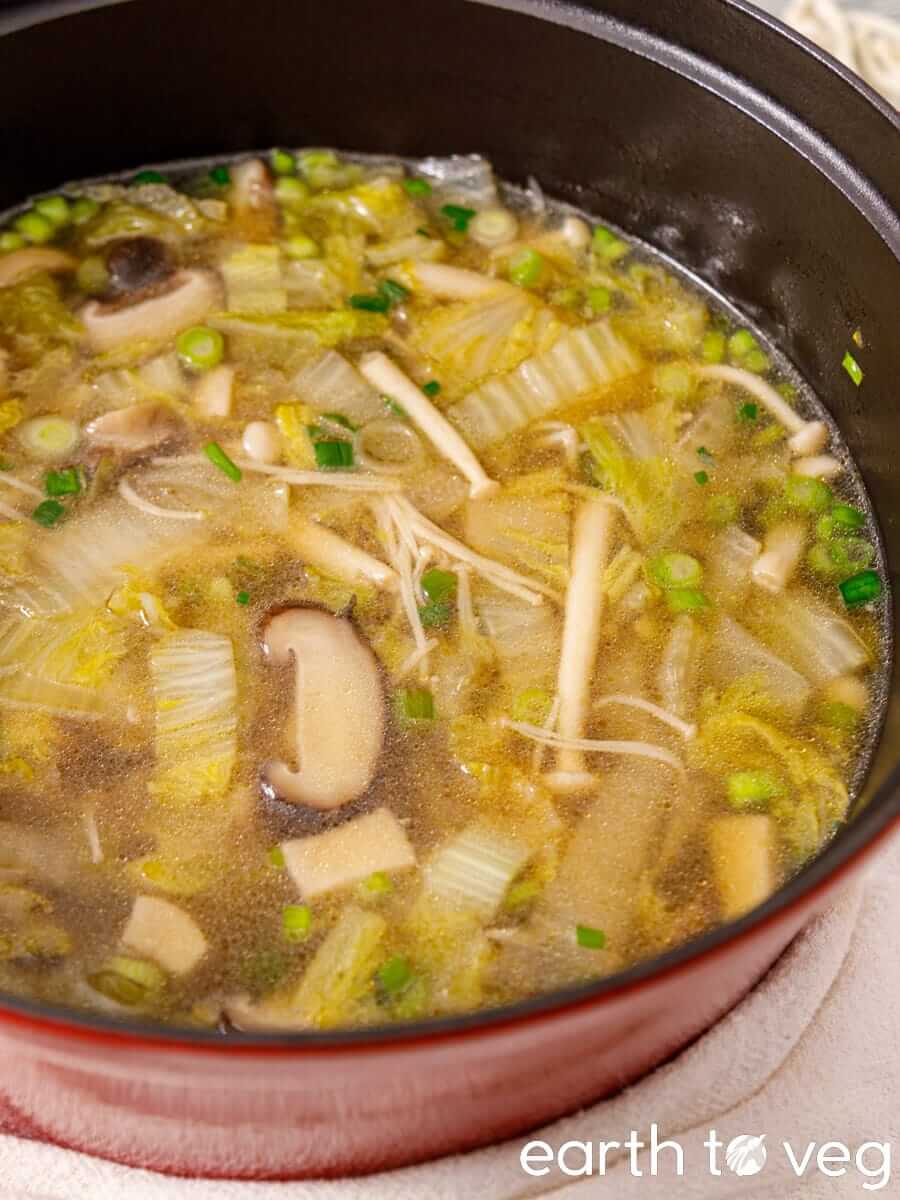 Soup in a red pot with cabbage, mixed mushrooms, and scallions in clear broth.